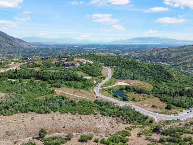 Expansive mountain landscape featuring a winding road, lush green foliage, distant urban development, a serene pond, and a broad blue sky with scattered clouds