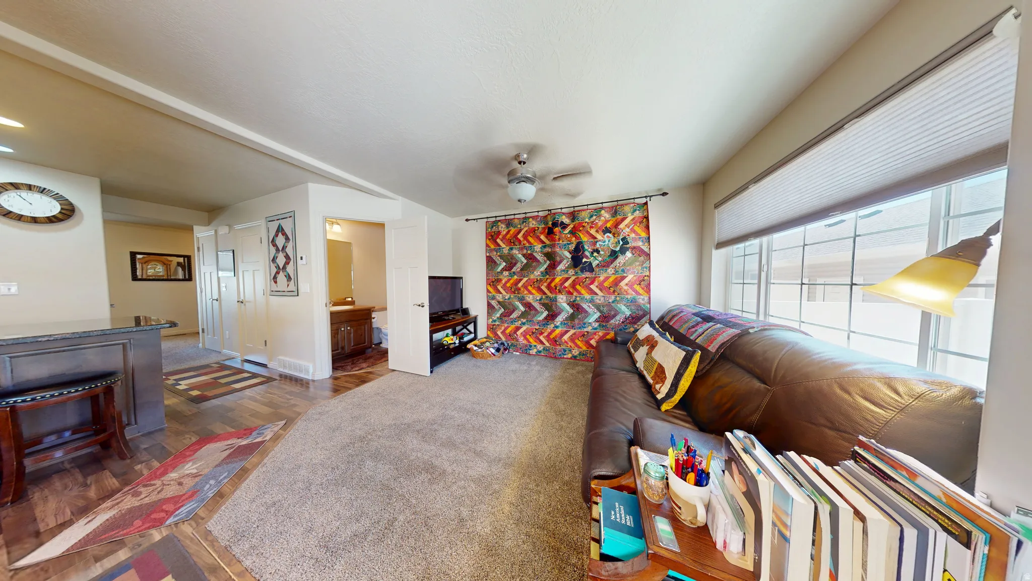 Living area featuring a textured carpet, ceiling fan, and large window with blinds