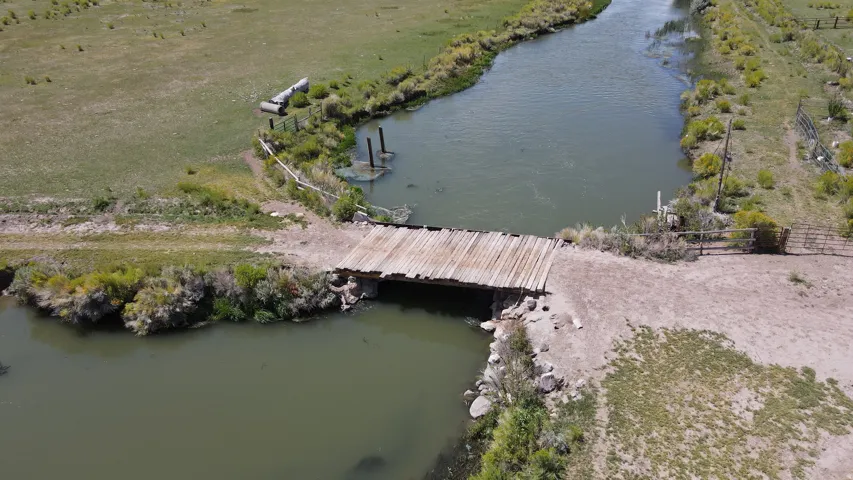 Waterway featuring a wooden bridge, grassy banks, and surrounding undeveloped land