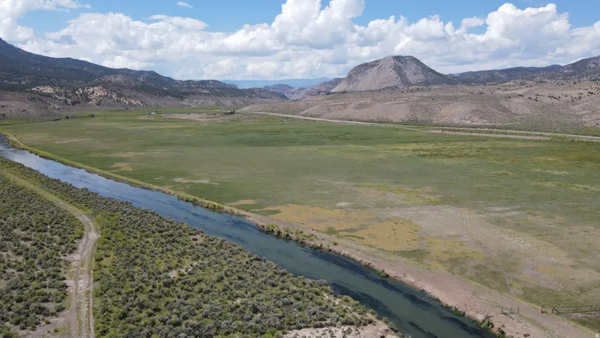 Expansive natural landscape featuring a prominent river, vast open fields, distant mountains, and a clear sky with scattered clouds