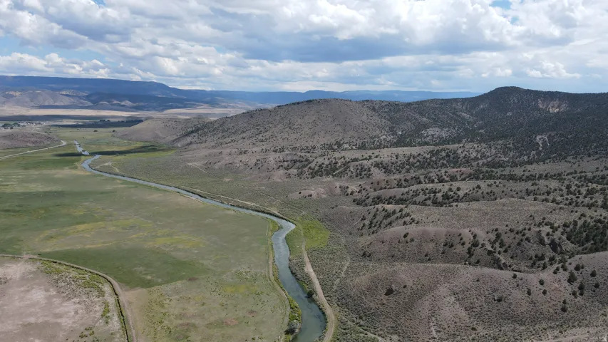 Expansive natural landscape featuring a winding river, verdant fields, and rugged, tree-dotted hillsides