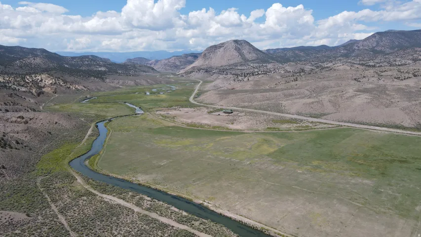Expansive land parcel featuring a river, verdant fields, a distant road, and surrounding mountainous terrain