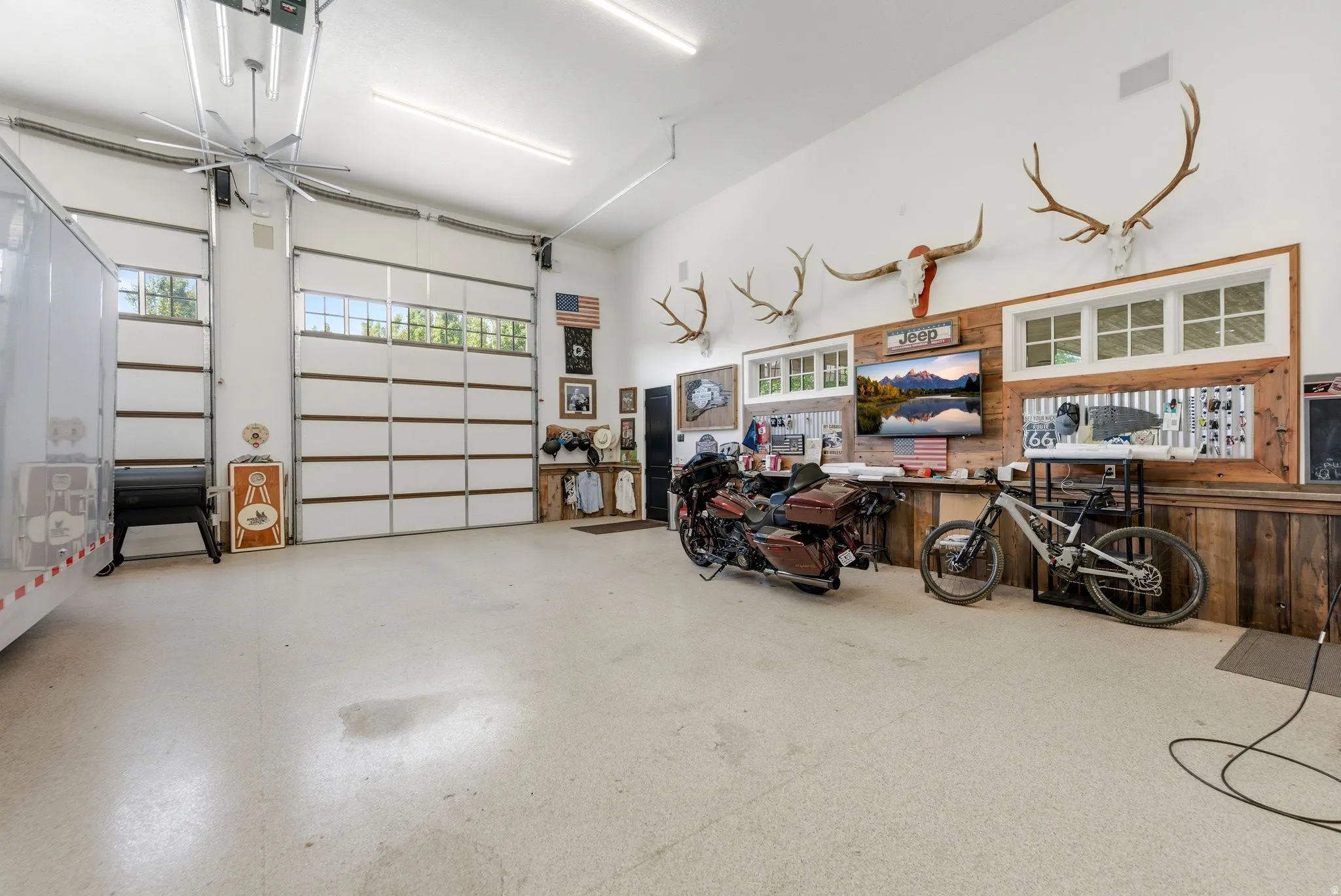 Spacious interior garage featuring two overhead doors with transom windows, epoxy flooring, a large ceiling fan, and extensive wall-mounted storage and display areas