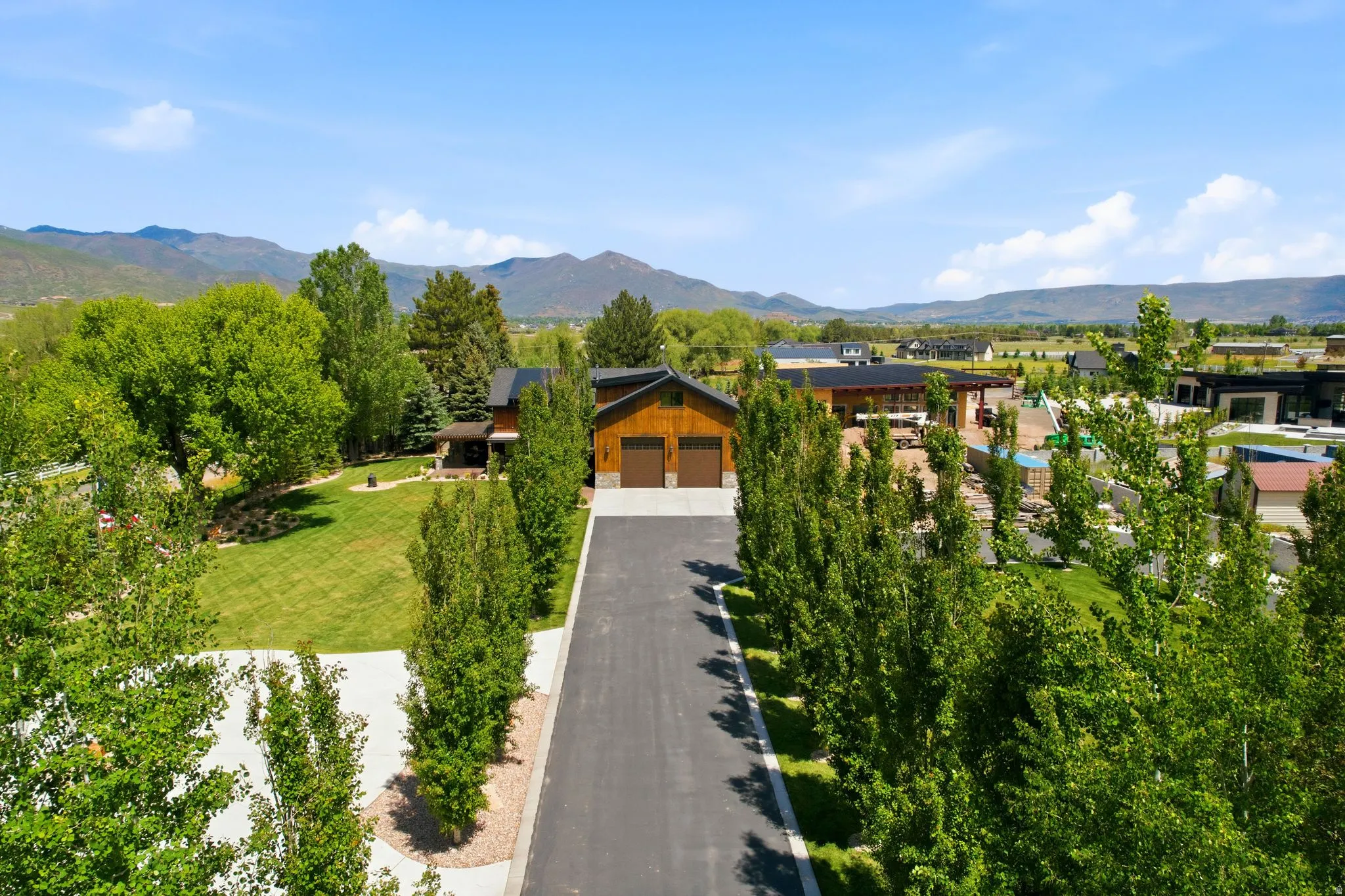 Expansive mountain landscape frames a property featuring a long paved driveway, a two-car garage with wood-paneled doors, and mature trees lining the perimeter