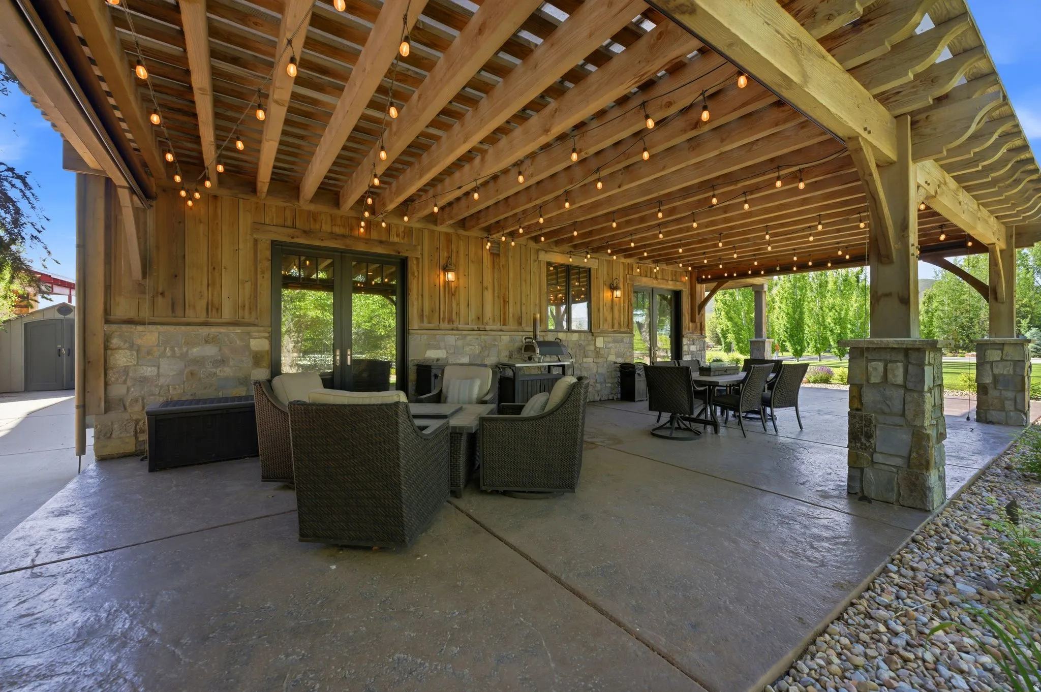 Expansive covered patio featuring a wood beam ceiling with string lighting, stone-clad pillars, and stamped concrete flooring