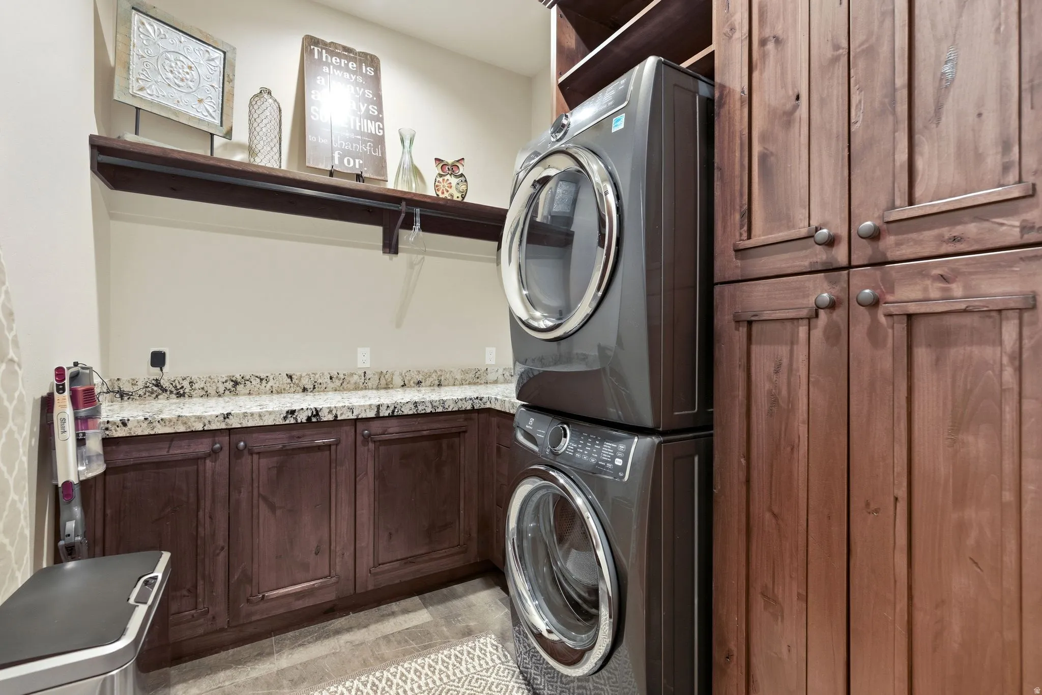 Laundry area featuring wood-finish cabinetry with silver hardware, light-toned granite-style countertops, and a dark wood floating shelf