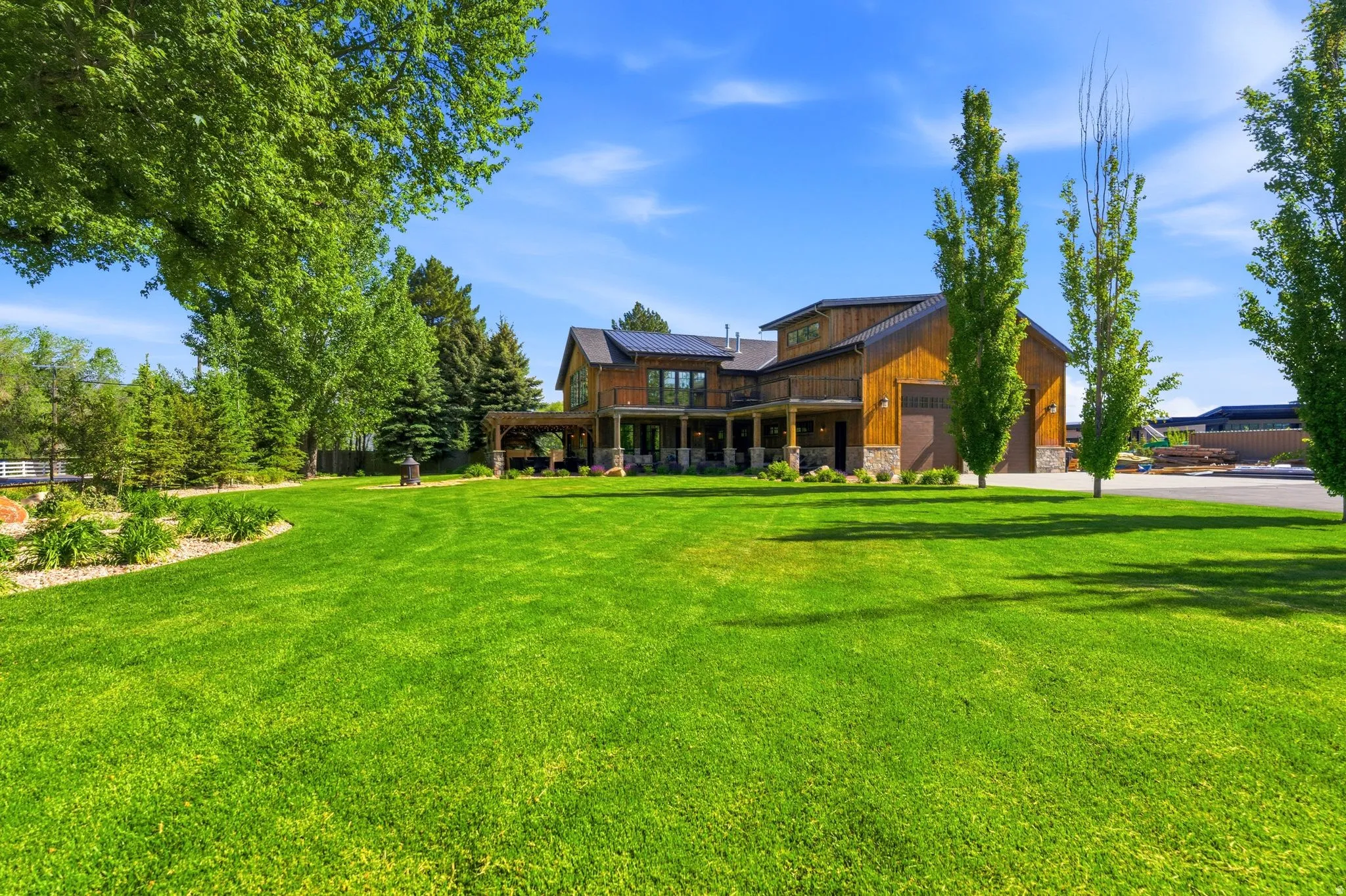 Expansive green lawn with mature trees bordering a multi-level wood-sided residence featuring a covered patio, stone column accents, and attached garage with two bay doors
