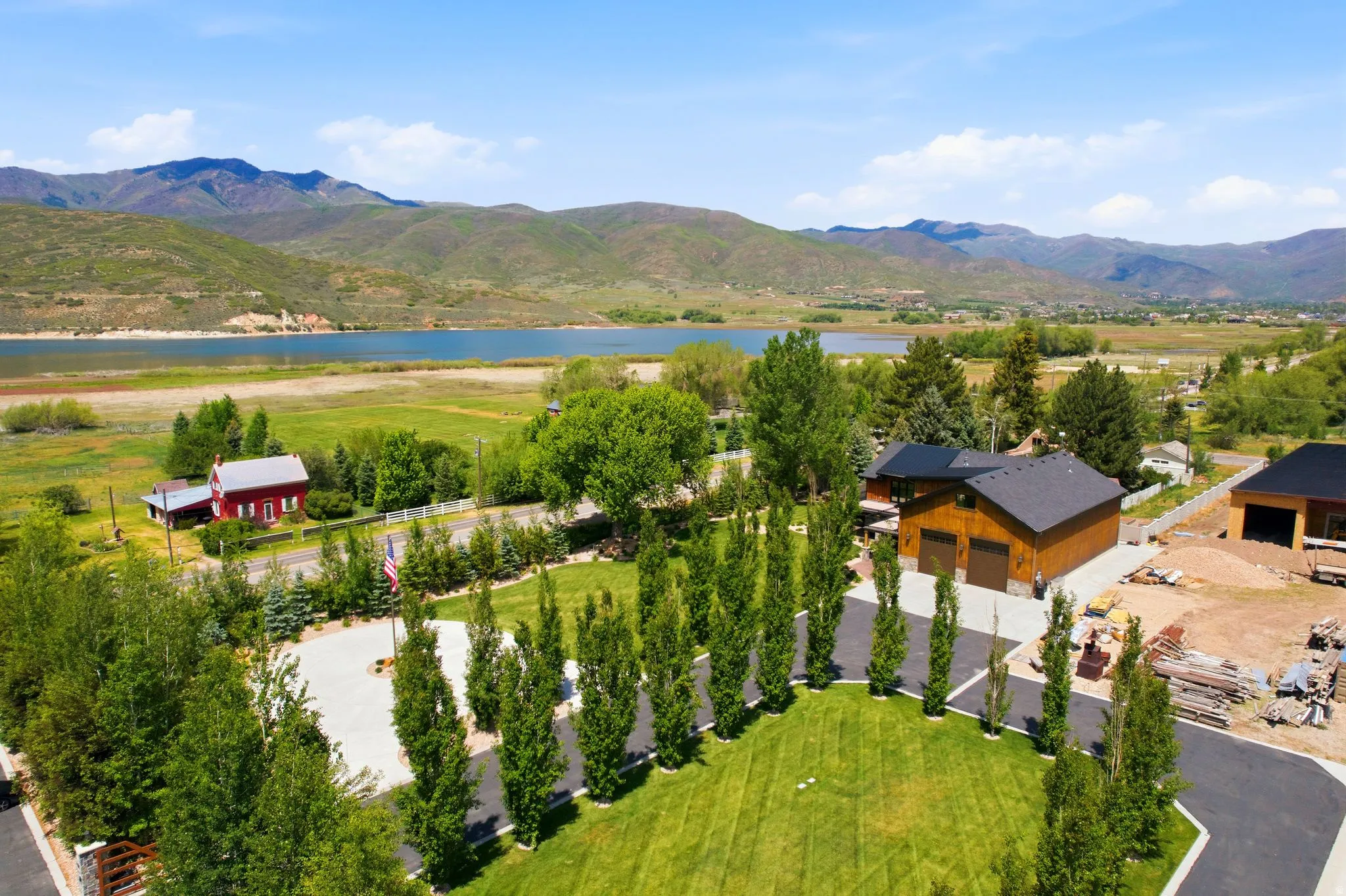 Expansive property featuring a paved circular driveway, mature landscaping, a dark-stained wood-sided home with a dark shingle roof, and a visible lake with mountainous terrain in the background