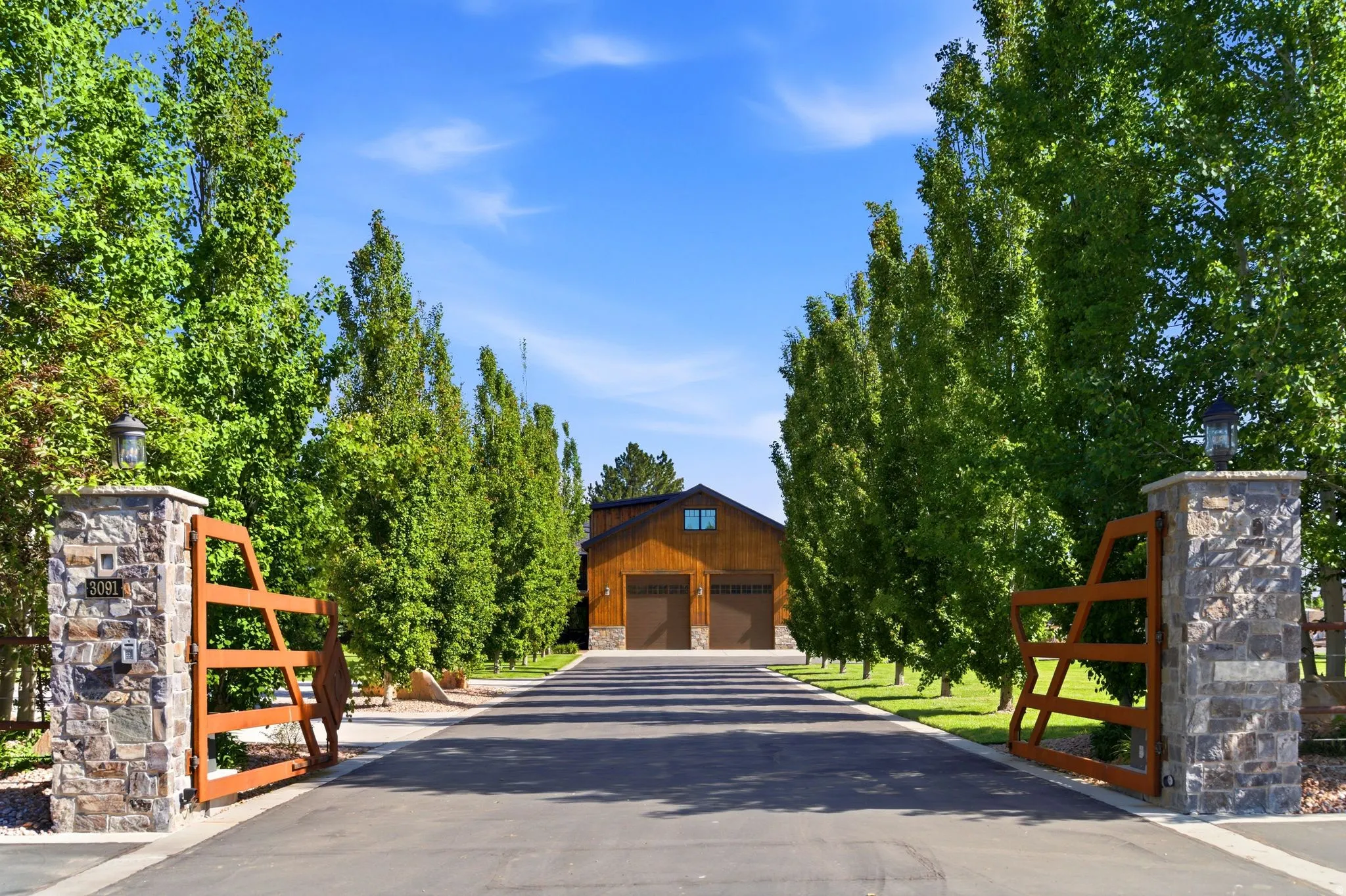 Paved driveway framed by mature trees, leading to a two-story structure featuring wood paneling, multiple garage doors, and upper-level windows