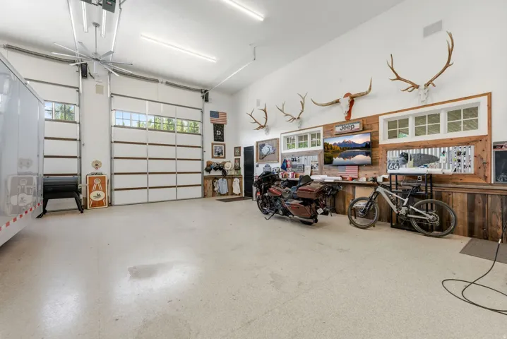 Spacious interior garage featuring two overhead doors with transom windows, epoxy flooring, a large ceiling fan, and extensive wall-mounted storage and display areas