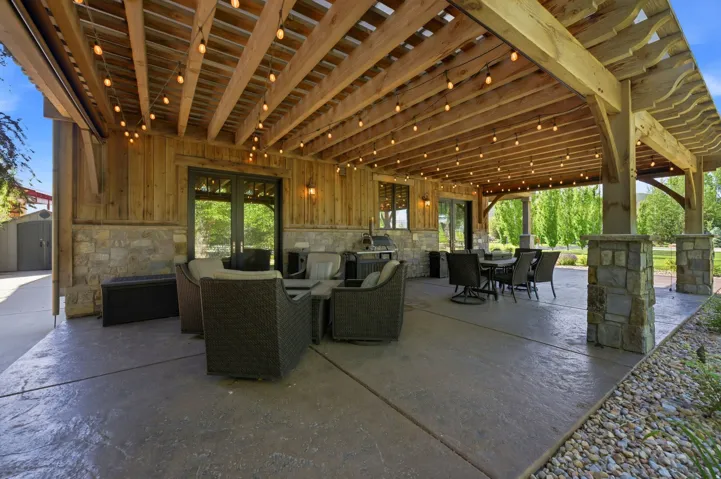 Expansive covered patio featuring a wood beam ceiling with string lighting, stone-clad pillars, and stamped concrete flooring