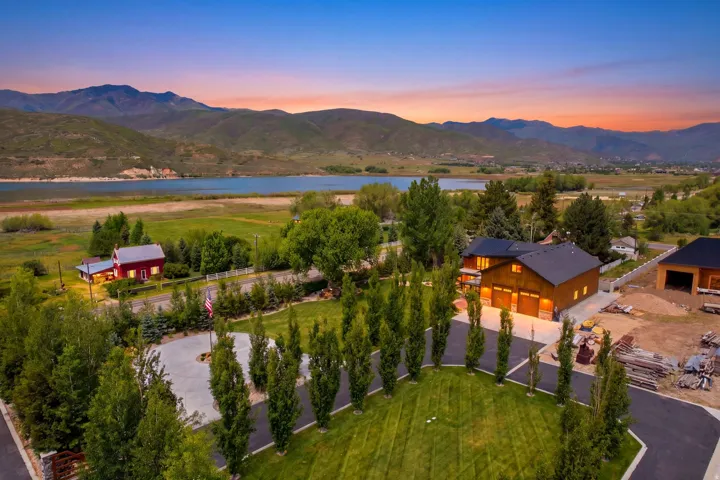 Expansive property featuring a wood-sided residence with an attached three-car garage, paved driveway, mature trees, and a mountain and lake backdrop