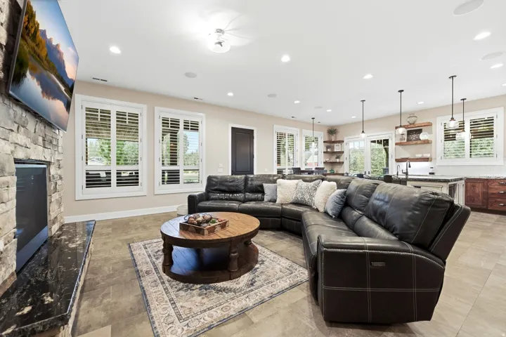 Spacious open-concept room featuring a stone-clad fireplace with a dark stone hearth, multiple windows with white plantation shutters, recessed ceiling lighting, and tile flooring