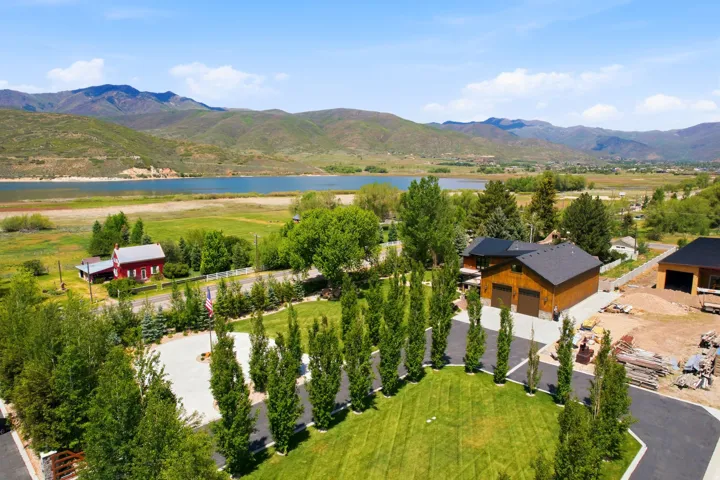 Expansive property featuring a paved circular driveway, mature landscaping, a dark-stained wood-sided home with a dark shingle roof, and a visible lake with mountainous terrain in the background