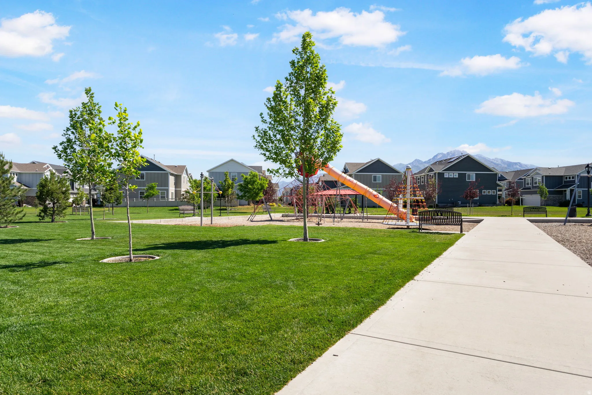 Spacious community green space featuring a playground, mature deciduous trees, and a paved pathway