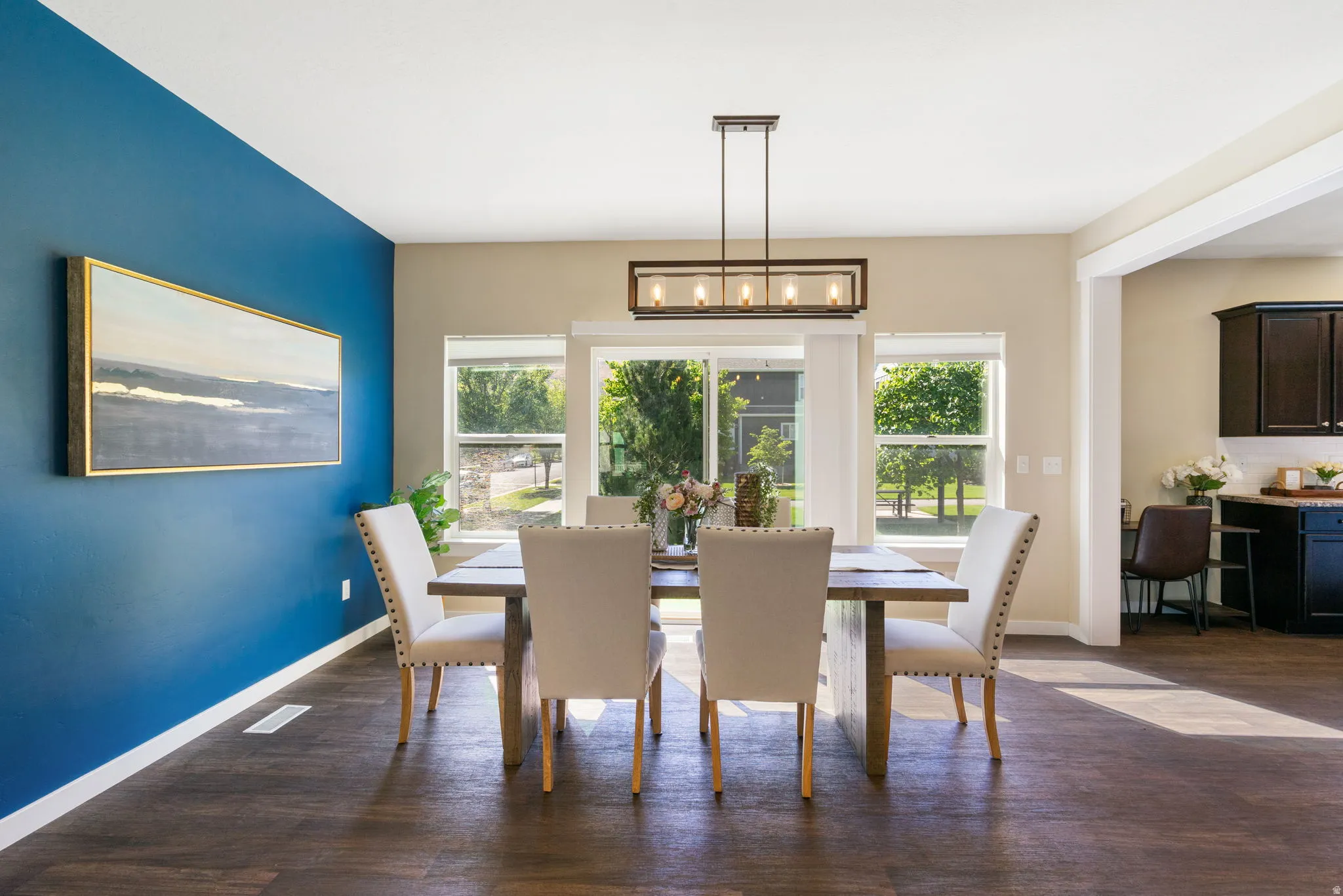 Dining area featuring wood-finish flooring, a linear chandelier, ample natural light from multiple windows, and a wide entryway to an adjacent kitchen space