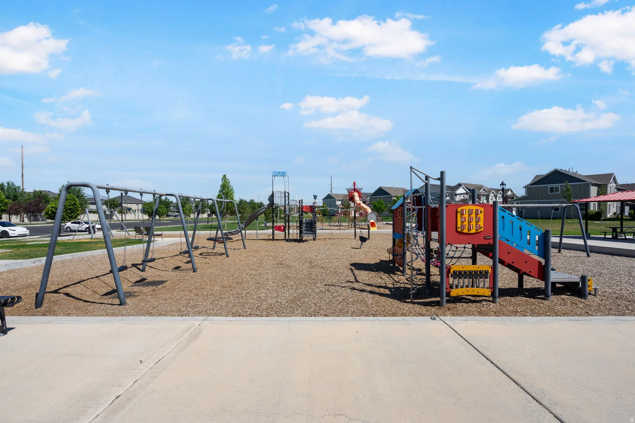 Community playground featuring multiple swing sets, a colorful play structure with slides, a climbing net, and wood chip surfacing