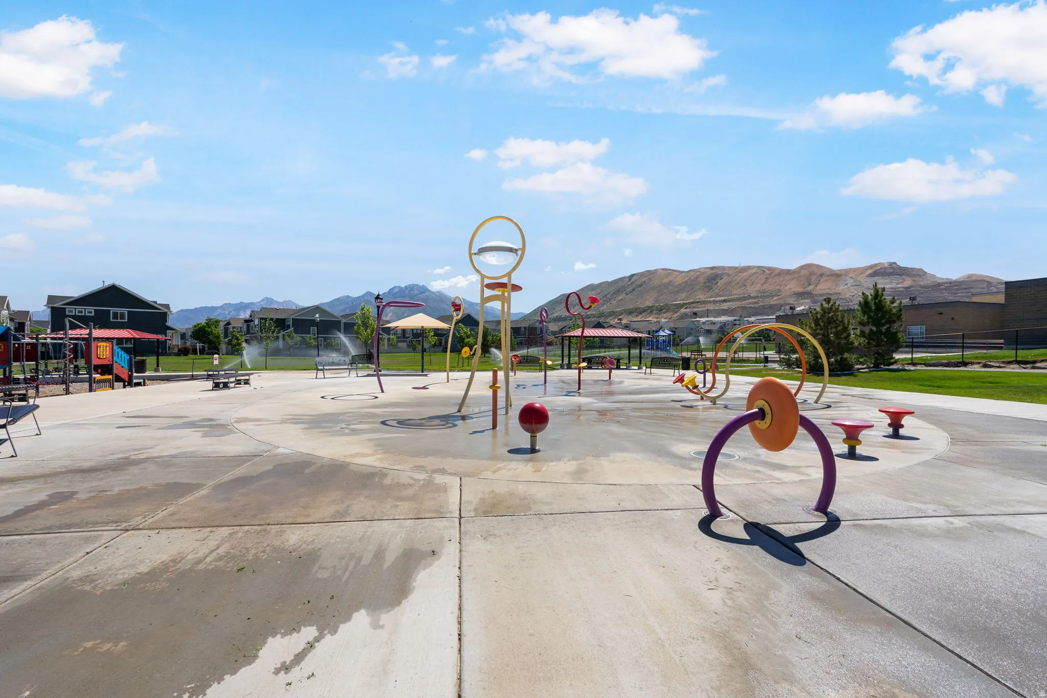 Community splash pad featuring multiple water play structures on a concrete surface, adjacent to green space with mountain scenery