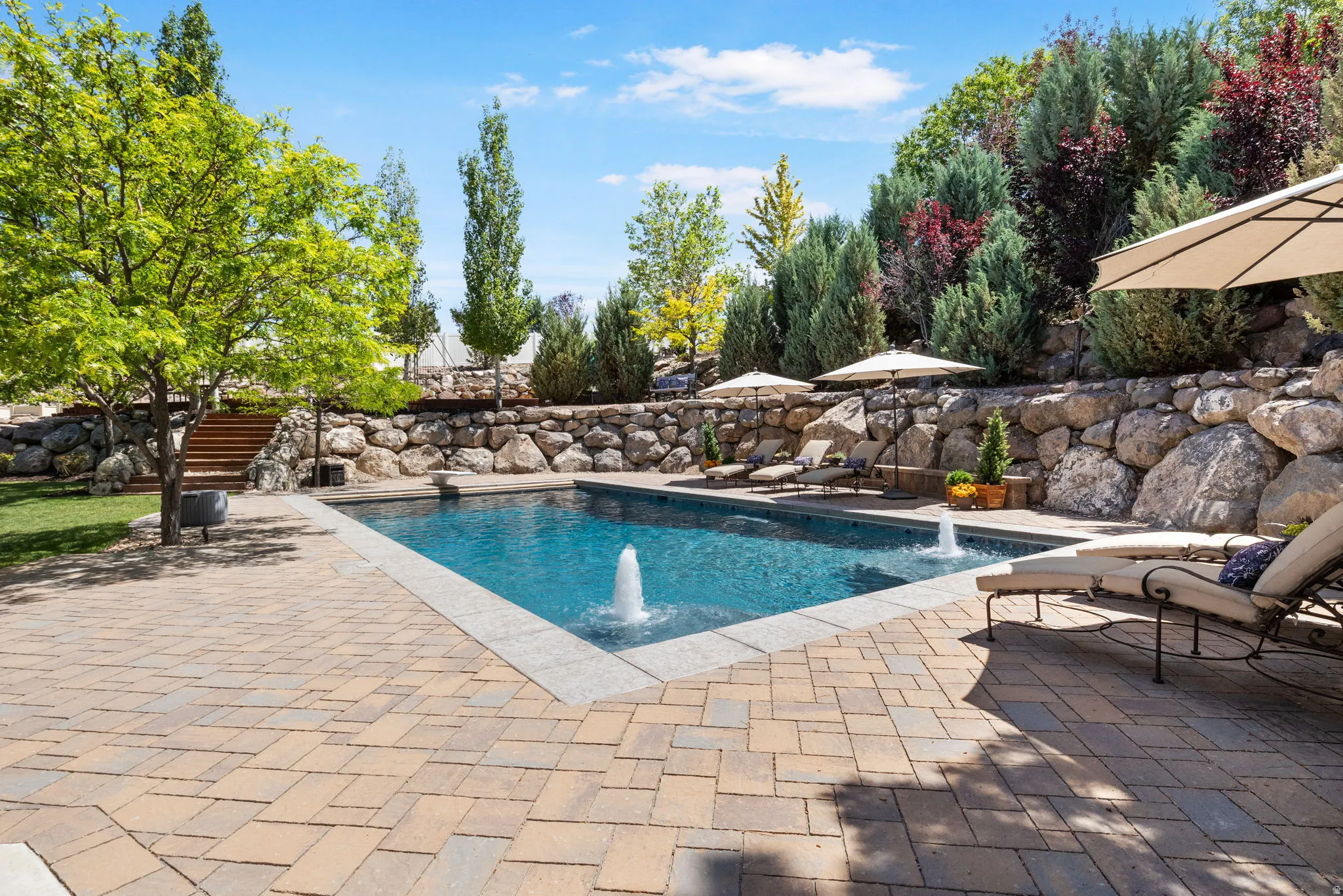 In-ground salt water swimming pool with bubblers, surrounded by a paver patio and a stone retaining wall with mature landscaping