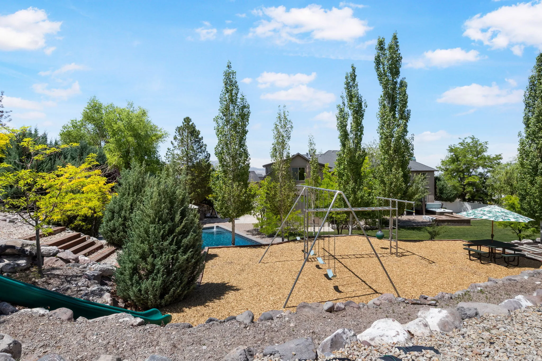 Landscaped backyard featuring a built-in swimming pool, a dedicated play area with wood chip surfacing, and mature trees providing natural screening