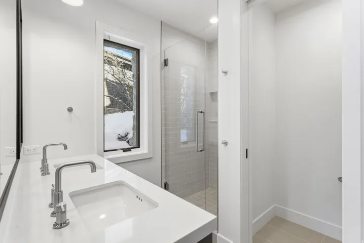 Bathroom featuring a dual vanity with integrated sinks and wall-mounted faucets, a frameless glass shower enclosure with subway tile surround, a tall vertical window, and light-toned flooring