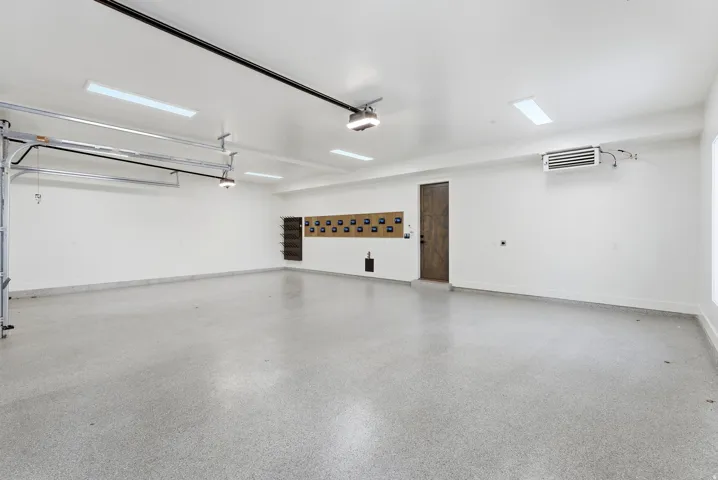 Spacious garage featuring epoxy flooring, crisp white walls, recessed fluorescent lighting, an industrial-style heater, and a dark wood-finish door
