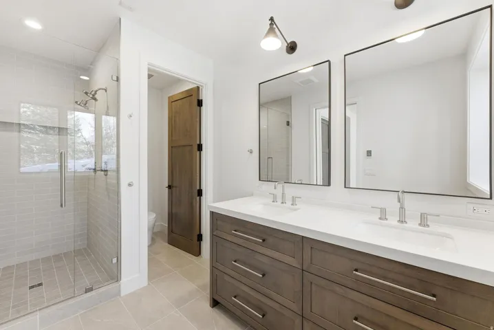 Bathroom featuring a glass-enclosed shower with subway tile surround, a dual vanity with a white countertop and wood-finish cabinetry, two rectangular framed mirrors, and light tile flooring
