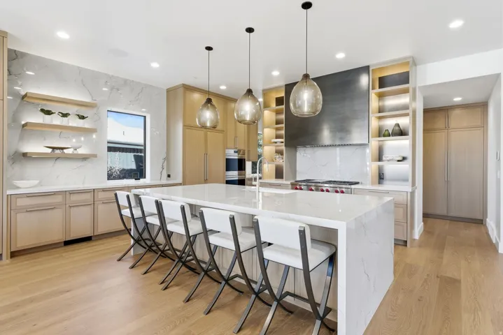 Expansive kitchen featuring a large central island with seating, light wood-finish cabinetry, integrated appliances, and a full-height stone-look backsplash