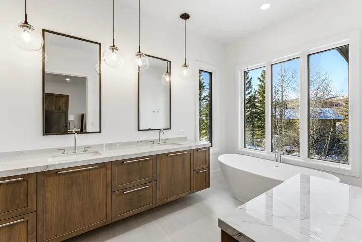 Modern bathroom featuring a freestanding tub, large windows, wood-finish double vanity with stone countertops, and pendant lighting