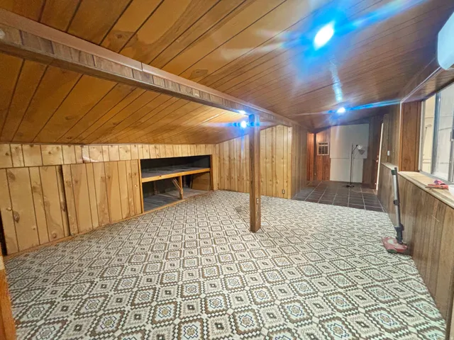 Spacious room featuring patterned carpet, wood paneling on walls and ceiling, built-in storage with shelving, and an adjacent tiled area