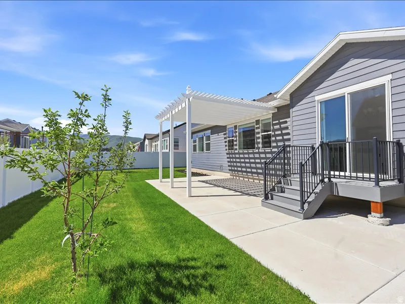 Backyard featuring a concrete patio with a white pergola, leading to a raised deck with dark railings
