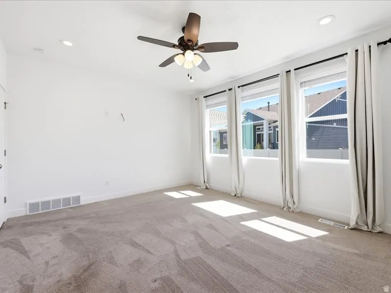 Spacious room featuring plush carpet flooring, a ceiling fan with integrated lighting, recessed lighting, and three large windows with curtain rods and drapes