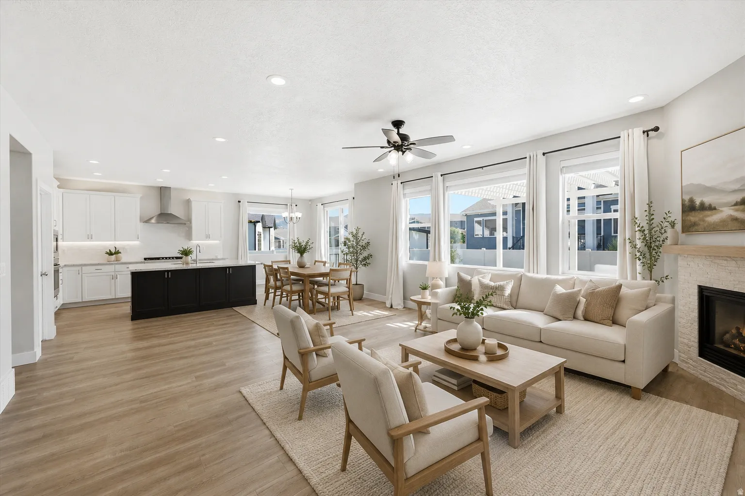 Open-concept living space featuring wood-finish flooring, a contemporary kitchen with white cabinetry and a dark island, a dining area with a chandelier, a living area with large windows, and a stone-clad fireplace