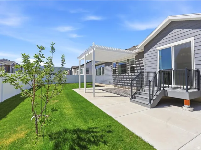 Backyard featuring a concrete patio with a white pergola, leading to a raised deck with dark railings