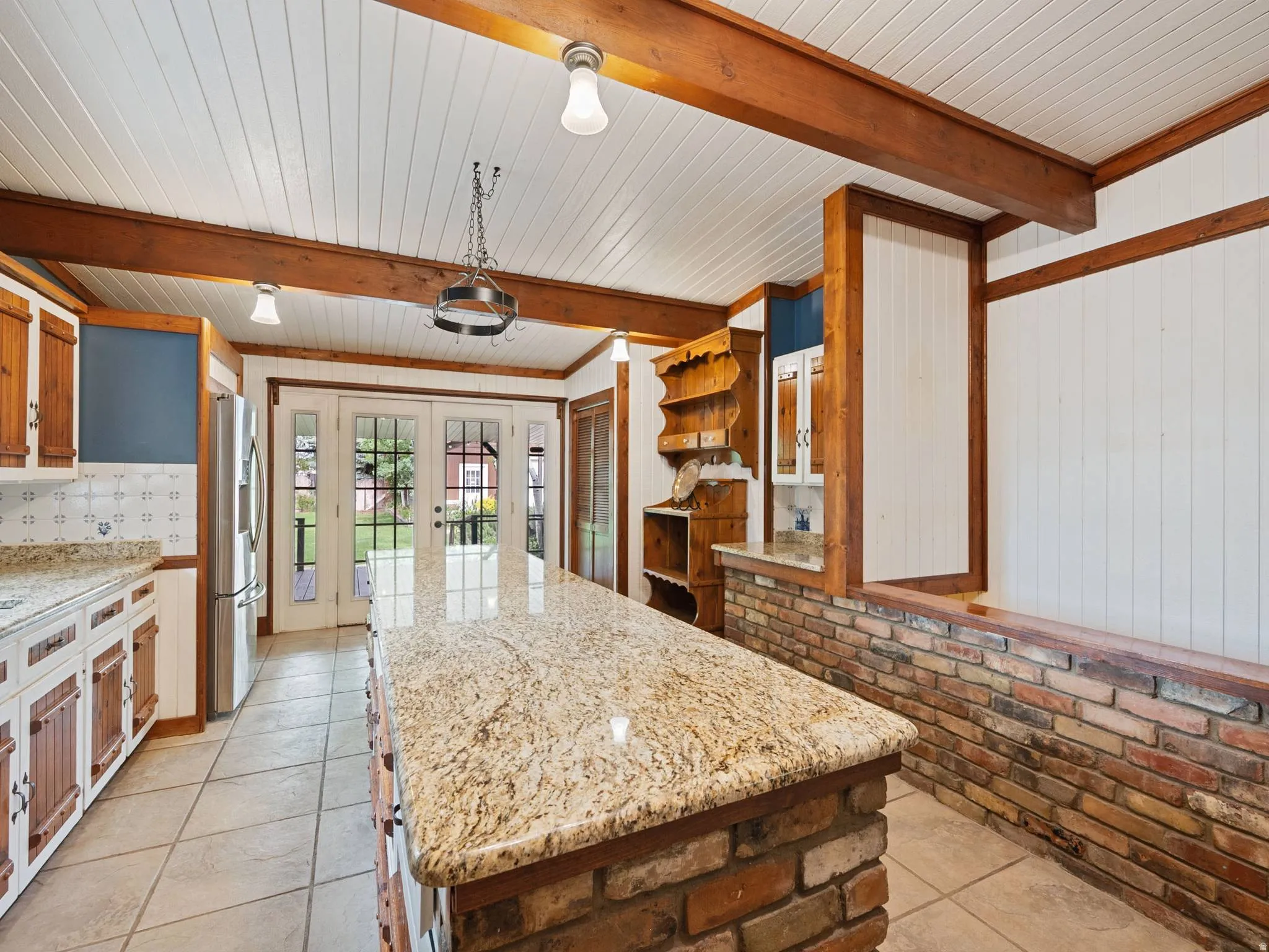 Large kitchen featuring a granite-topped island, exposed wood beams, brick accents, and French doors leading to the backyard area.