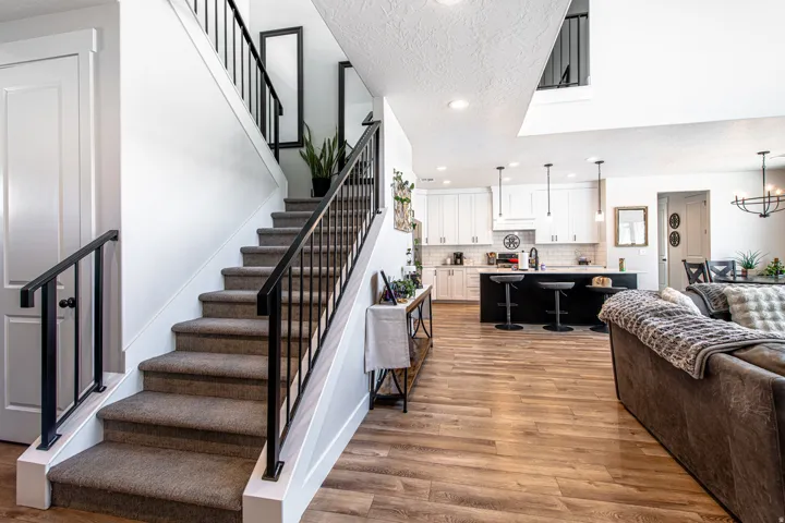 Open-concept interior featuring wood-finish flooring, a carpeted staircase with black metal railings, white shaker-style cabinetry, a contrasting black kitchen island with pendant lighting, and recessed ceiling lights