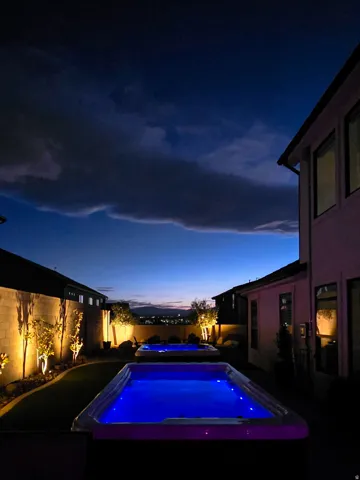 Backyard oasis featuring two illuminated pools, artificial turf, and mature landscaping with uplighting