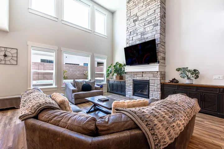 Two-story living area featuring a stacked stone fireplace with a white mantel, wood-finish flooring, and multiple windows including upper transom windows