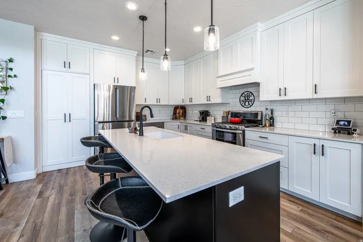 Contemporary kitchen featuring an expansive island with a light-toned countertop, white shaker cabinetry, stainless steel appliances, a subway tile backsplash, and wood-finish flooring