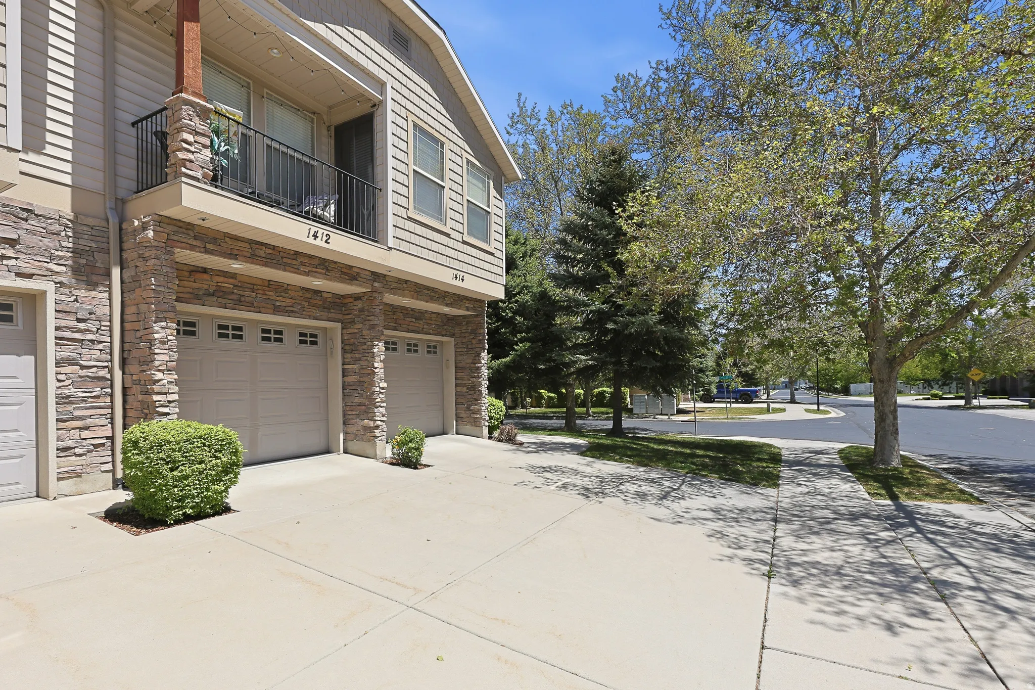 Exterior featuring stacked stone accents, horizontal siding, private balcony with black iron railing, two garage doors, and mature trees lining the street