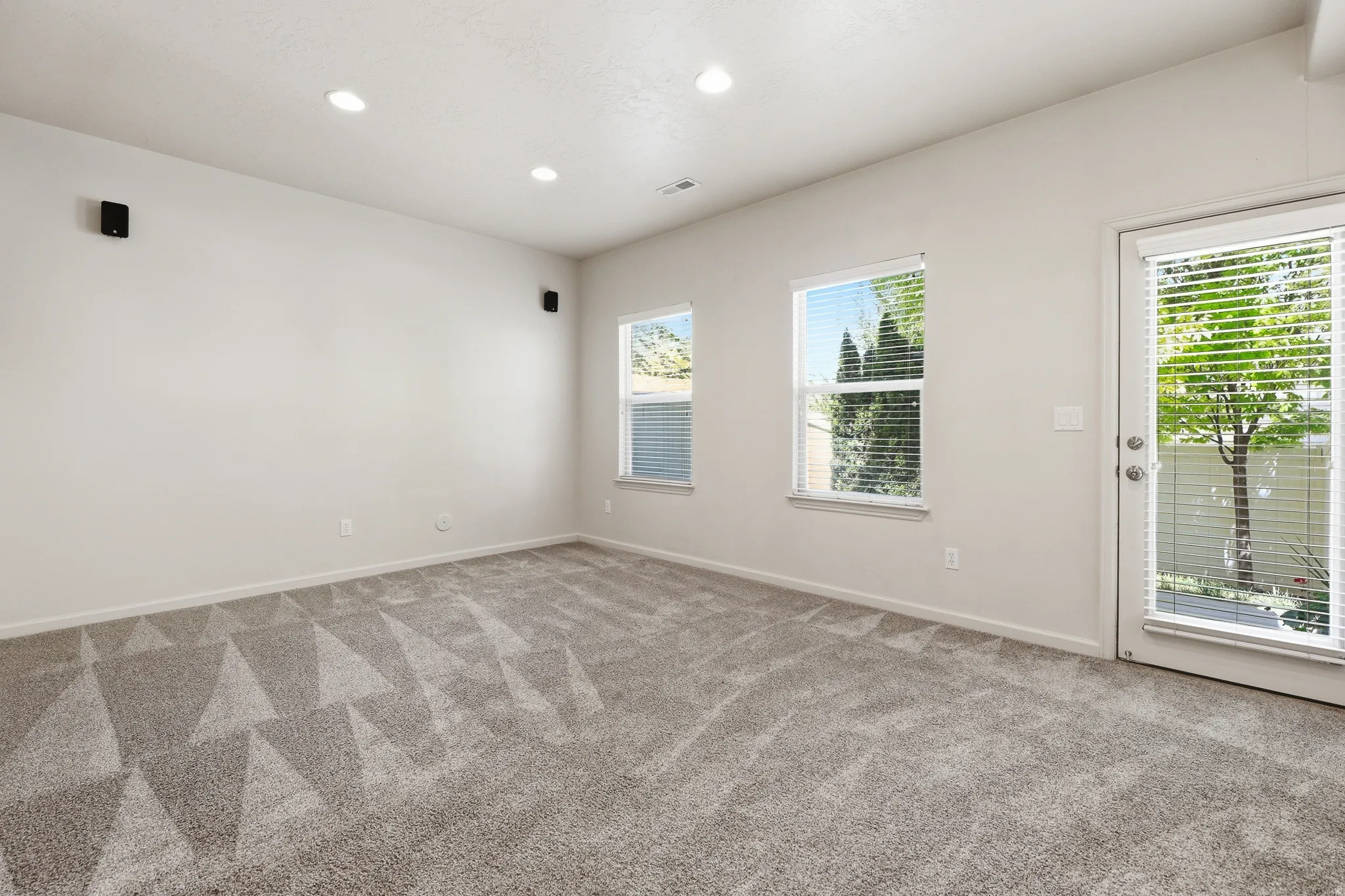 Spacious room featuring neutral carpeting with a geometric pattern, recessed lighting, and wall-mounted speakers