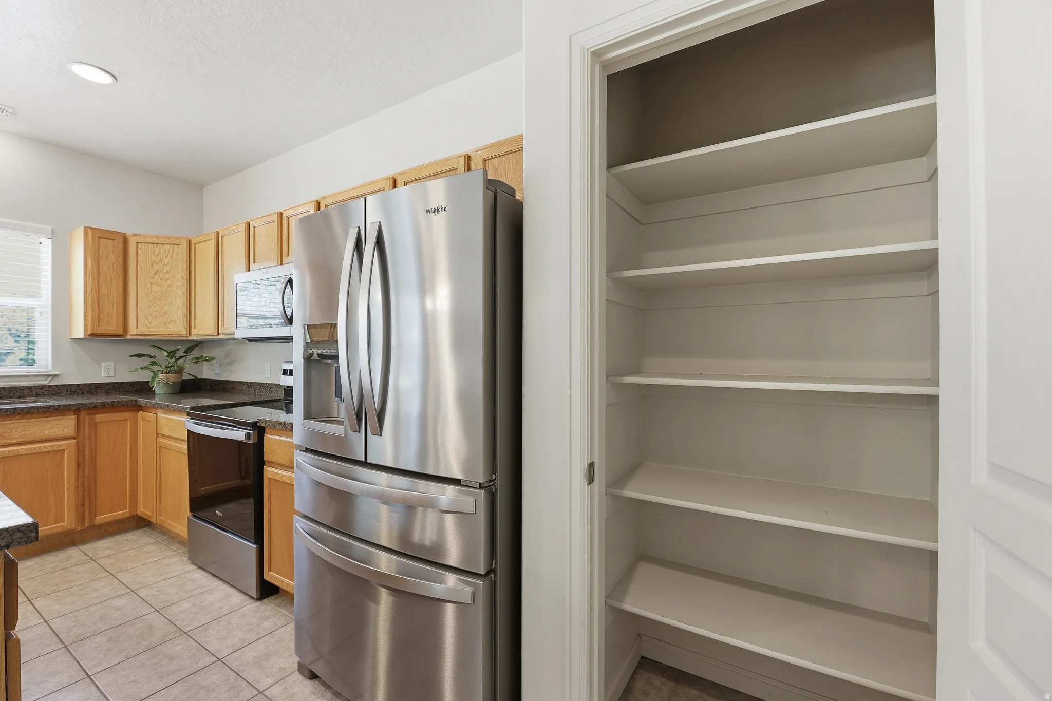 Kitchen pantry with multiple fixed shelves, alongside a kitchen featuring light wood cabinetry, stone-finish countertops, stainless steel appliances, and tile flooring