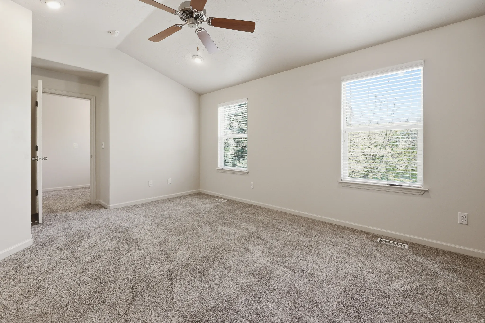 Spacious room featuring vaulted ceilings, recessed lighting, a ceiling fan with wood-finish blades, soft grey carpeting, and two windows with blinds