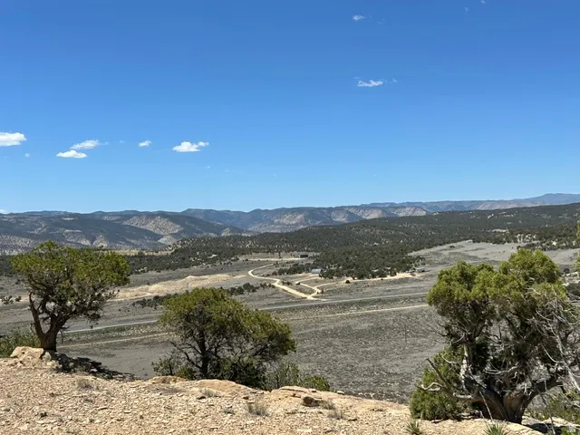 Expansive mountain desert landscape featuring rugged terrain, distant mountain ranges, scattered green vegetation, and a clear blue sky