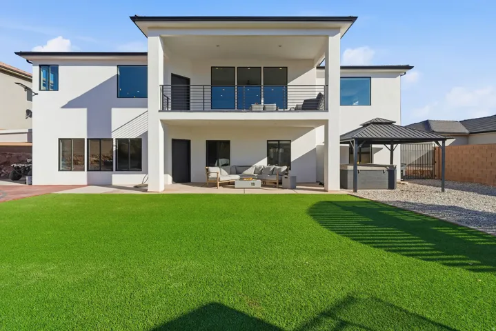 Contemporary two-story exterior featuring a ground-level covered patio, an upper-level covered balcony with a metal railing, extensive green lawn, a detached gazebo, and a gravel-covered area