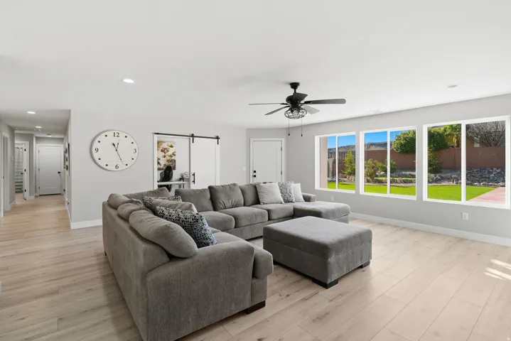 Spacious living area featuring light wood-finish flooring, a contemporary ceiling fan, recessed lighting, and multiple large windows overlooking a landscaped backyard