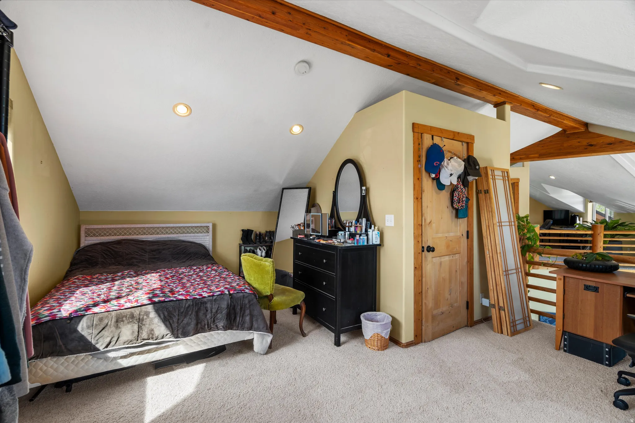 Carpeted room featuring a white headboard, recessed lighting, exposed ceiling beams, a natural wood paneled door, and a wooden railing