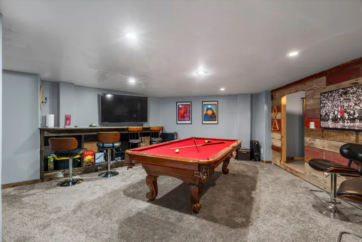 Spacious entertainment room featuring recessed lighting, gray carpet flooring, a built-in bar with wood-finish paneling, and a rustic wood-finish accent wall