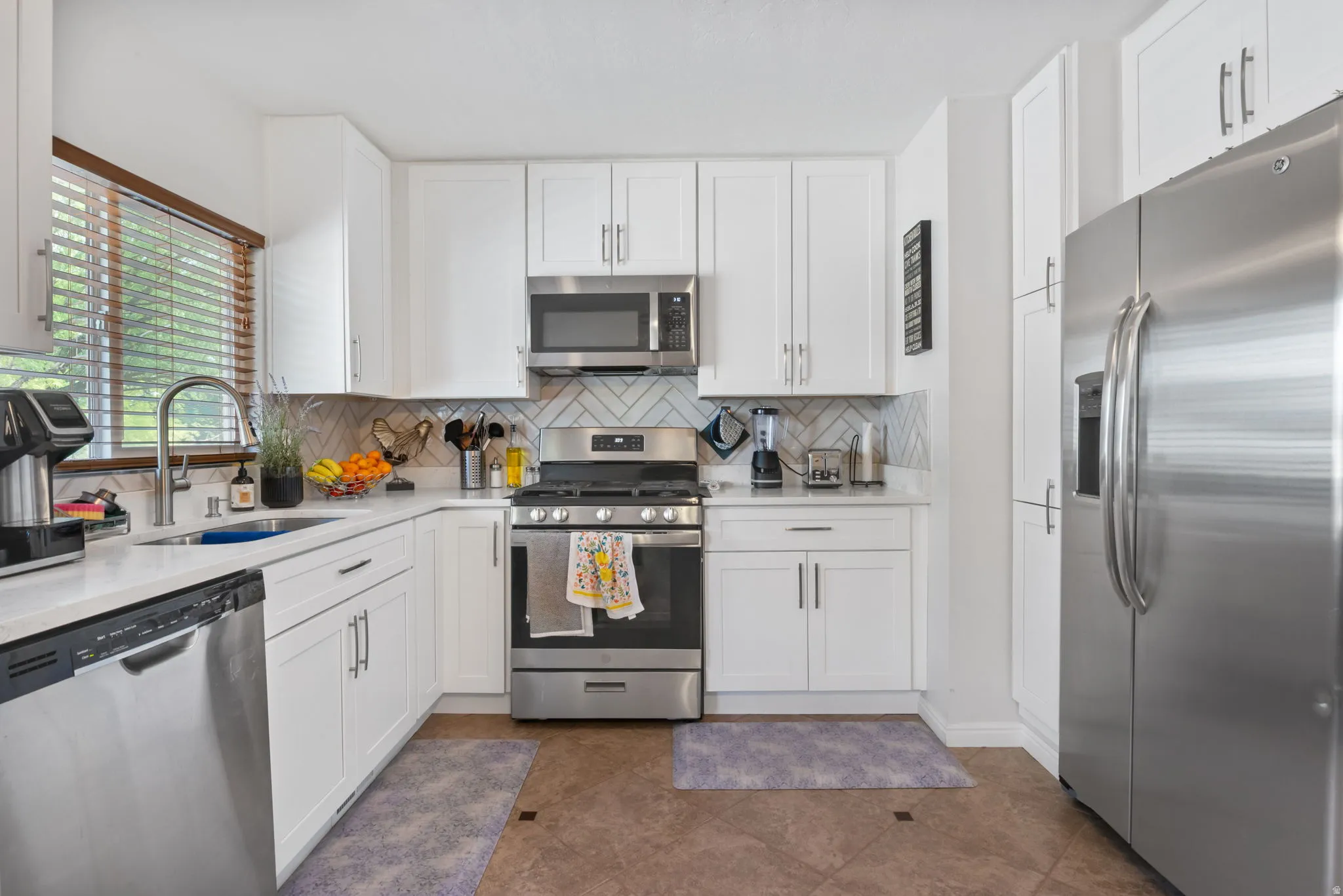 Modern kitchen featuring white shaker cabinetry, stainless steel appliances, white countertops, a herringbone tile backsplash, and wood-finish blinds