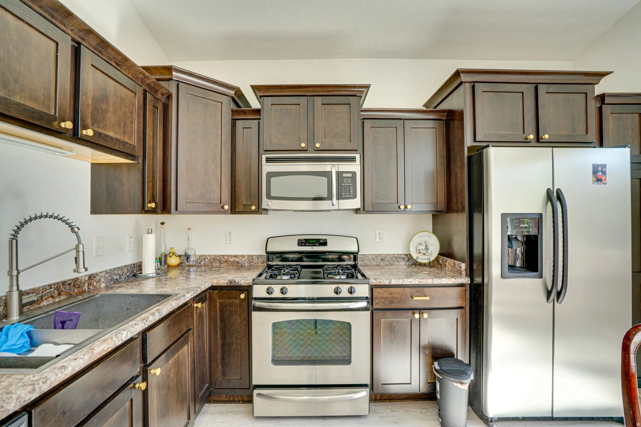 Kitchen featuring dark wood-finish cabinetry with gold-tone hardware, stainless steel appliances, a double basin sink with a high-arc faucet, and granite-look countertops